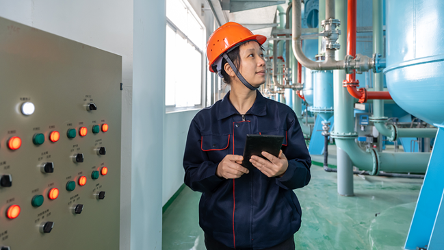 Auf dem Bild zu sehen ist eine Ingenieurin die einen Tablet-Computer nutzt, um Geräte in einer Fabrik zu überprüfen. Auf dem Bild zu sehen ist eine Ingenieurin die einen Tablet-Computer nutzt, um Geräte in einer Fabrik zu überprüfen.